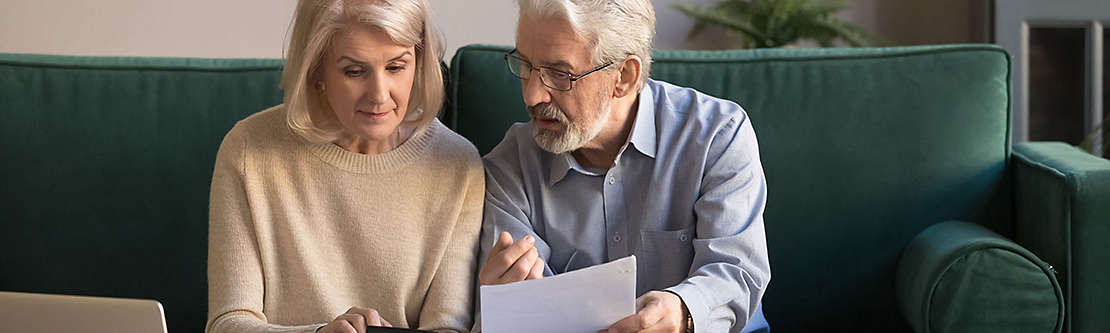 A mature couple siting on the couch looking a paperwork. 