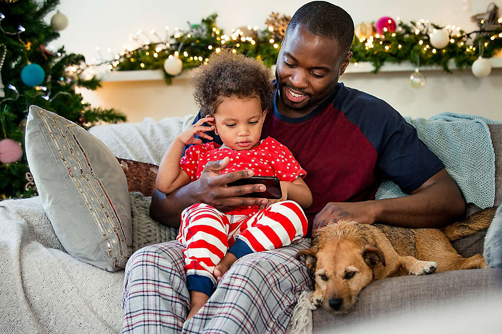 A father and daughter sitting on the couch with their dog, looking at his phone.