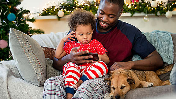 A father and daughter sitting on the couch with their dog, looking at his phone.