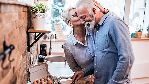 A mature woman kissing a mature man while he's cooking breakfast