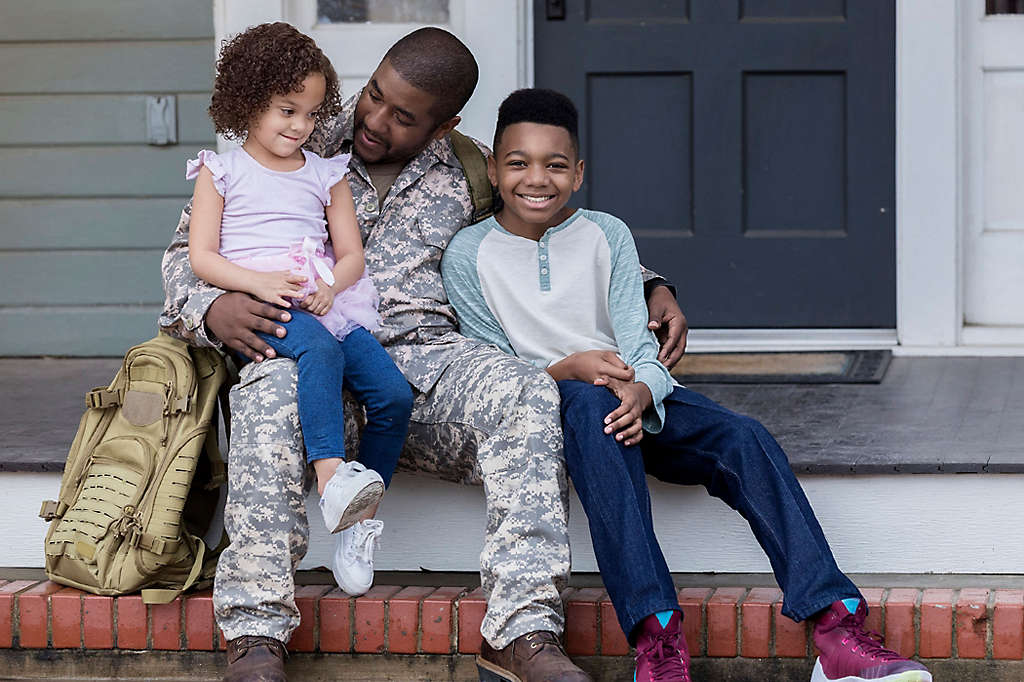 An active duty serviceman in uniform sitting on the porch hugging his children.