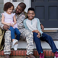 An active duty serviceman in uniform sitting on the porch hugging his children.