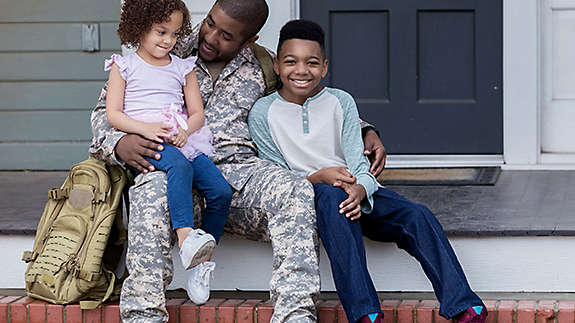 An active duty serviceman in uniform sitting on the porch hugging his children.