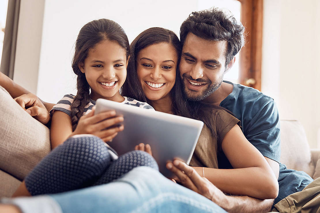 A family sitting on the couch happily looking at something on a tablet together.