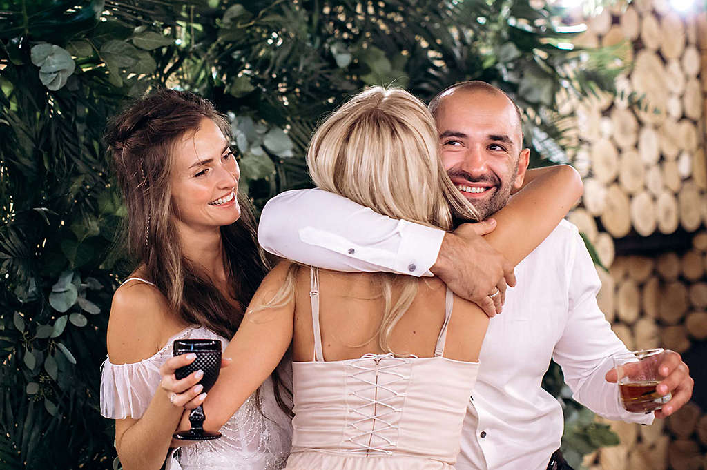 Woman hugging couple while holding drinks and celebrating.