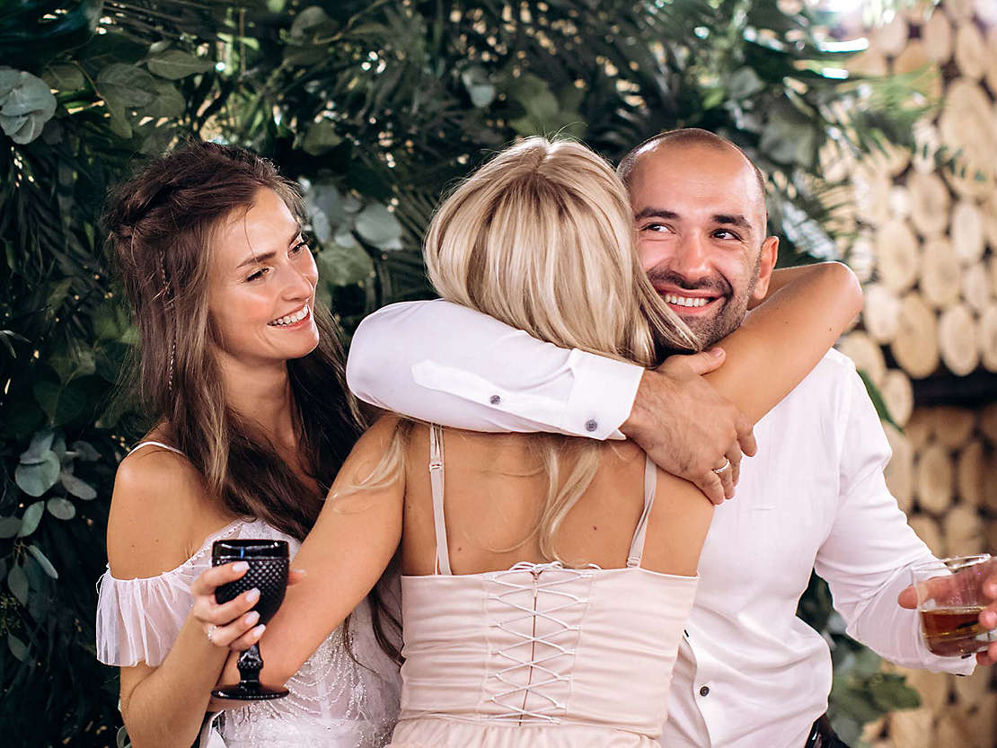 Woman hugging couple while holding drinks and celebrating.
