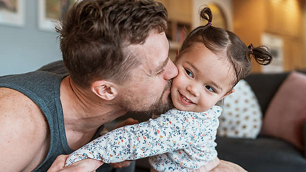 Father kissing young daughter on the cheek while sitting on a living room couch