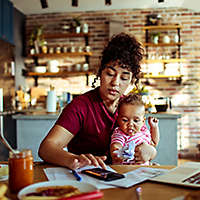A young mother using a calculator while sitting at a table holding her baby.