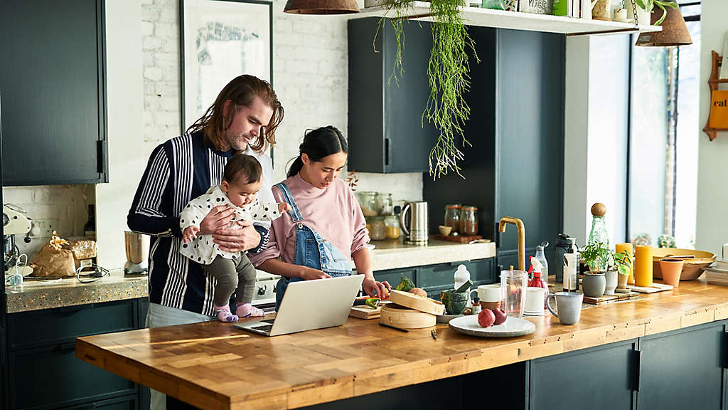 Two parents with their baby daughter using computer and making dinner at home, domestic life, technology, routine