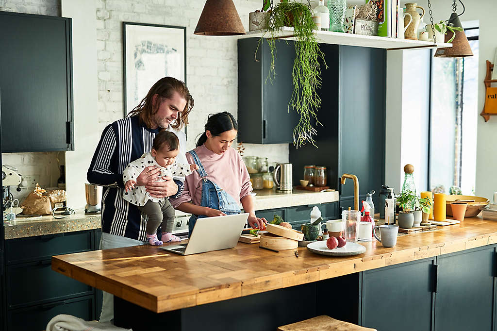 Two parents with their baby daughter using computer and making dinner at home, domestic life, technology, routine