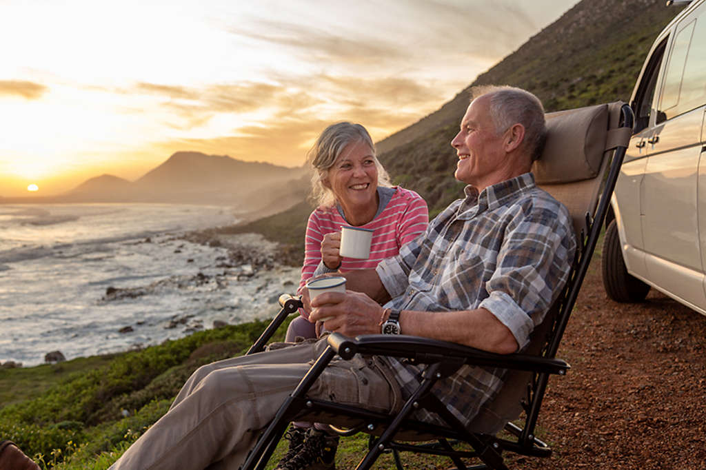 Retired couple sitting on lounge chairs on a seaside cliff at sunrise drinking coffee and laughing together