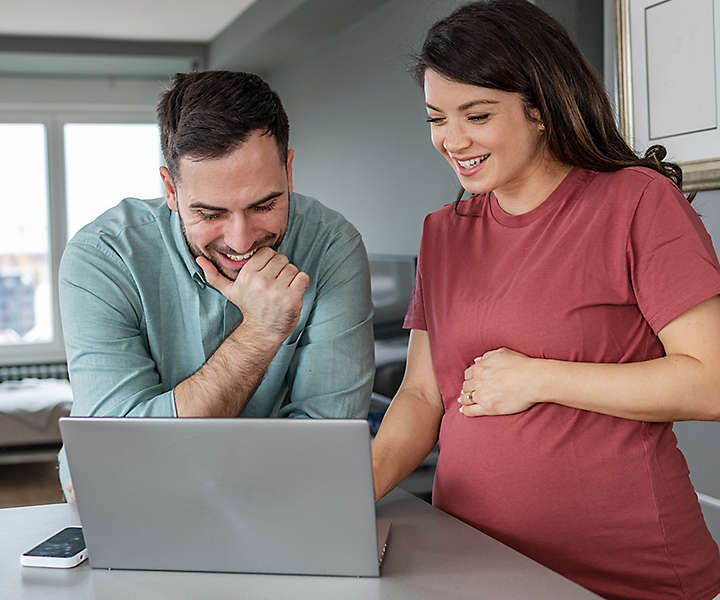 Lovely pregnant couple watching videos about pregnancy on the internet. They are smiling while looking at the laptop. Preparation for parenting.