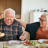 Elderly couple laughing and eating lunch in their kitchen.