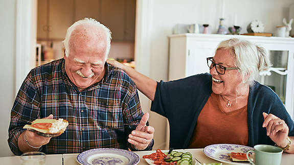 Elderly couple laughing and eating lunch in their kitchen.