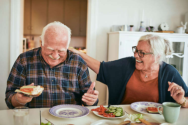 Elderly couple laughing and eating lunch in their kitchen.