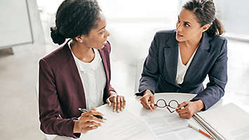 Two women sitting at a table talking and filling out paperwork.