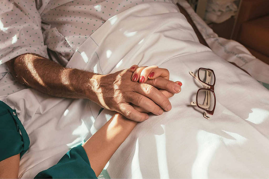 A daughter holds her father’s hand as he lies in a hospital bed.
