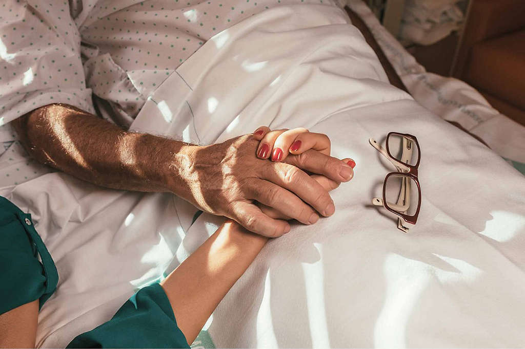 A daughter holds her father’s hand as he lies in a hospital bed. 