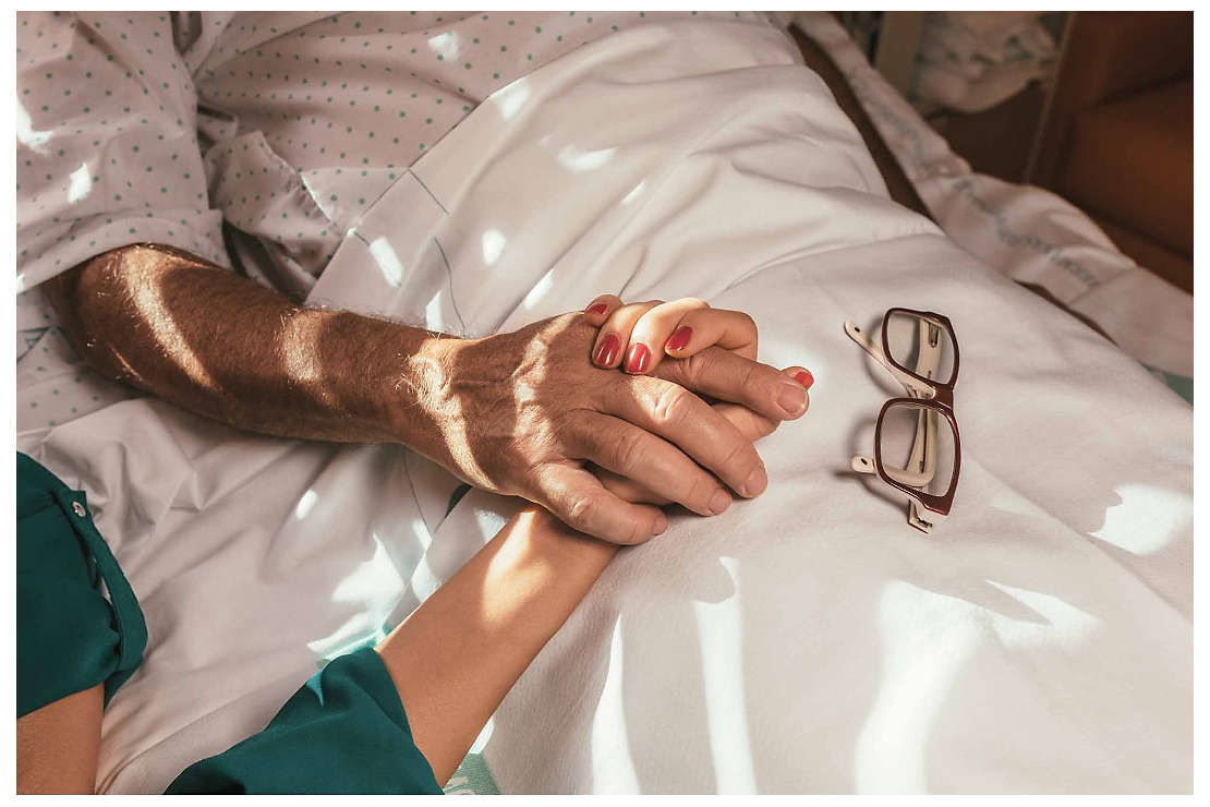 A daughter holds her father’s hand as he lies in a hospital bed.