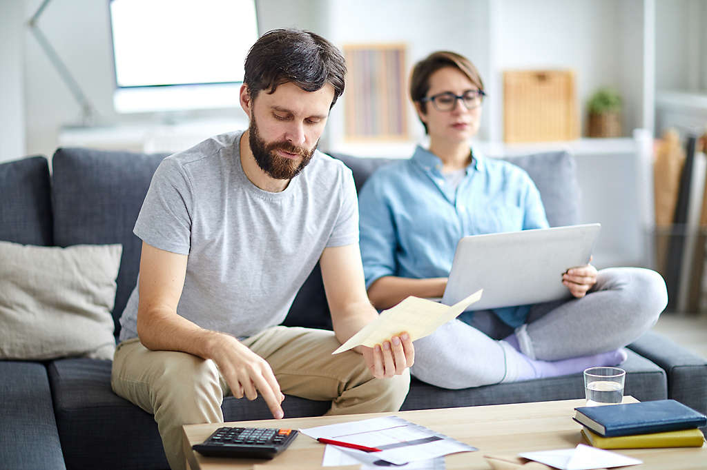 A married couple sitting on a couch, looking through bills, and working on a computer.