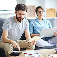 A married couple sitting on a couch, looking through bills, and working on a computer.