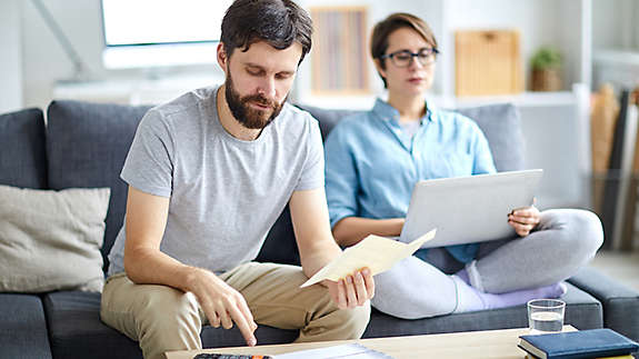 A married couple sitting on a couch, looking through bills, and working on a computer.