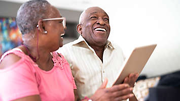 A mature couple laughing and smiling at something on a tablet.