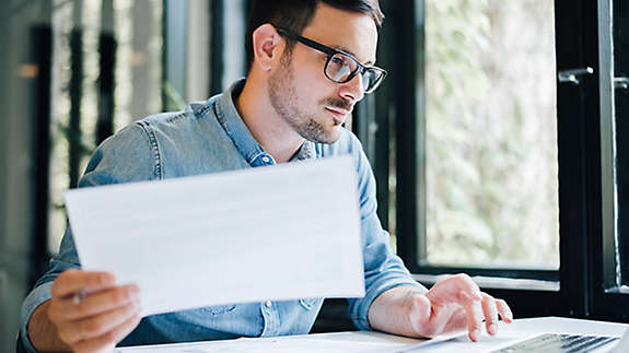 A man sitting at a desk holding paperwork and working on a computer.