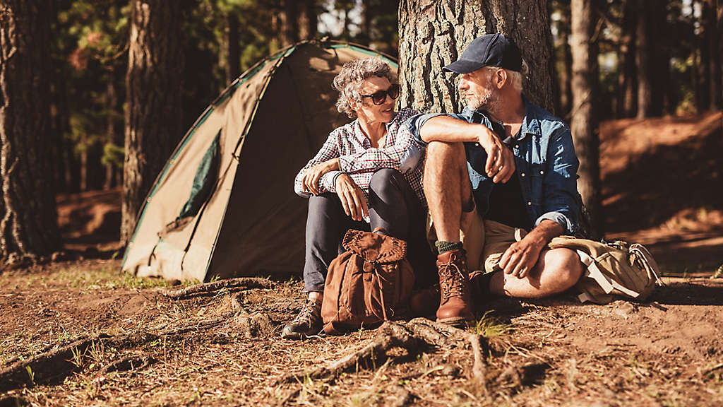 A mature couple sitting under a tree outside a camping tent.