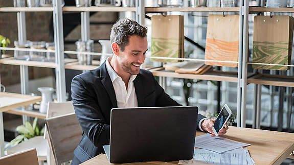 A man at a table working on a laptop and looking at his cell phone.