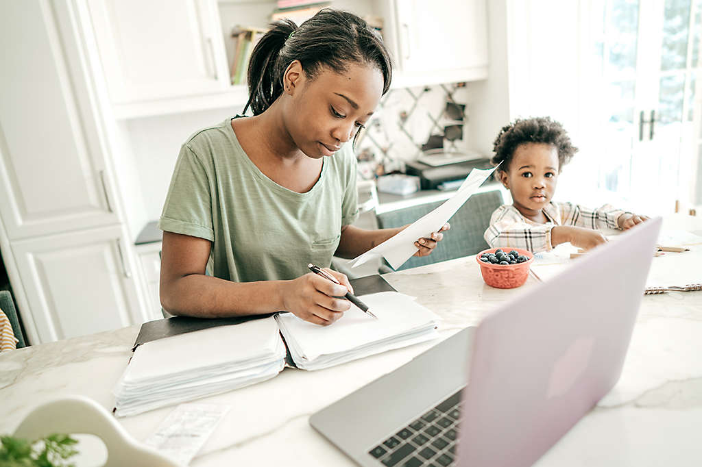 A woman sitting at a table reviewing paperwork while her daughter draws on a tablet.