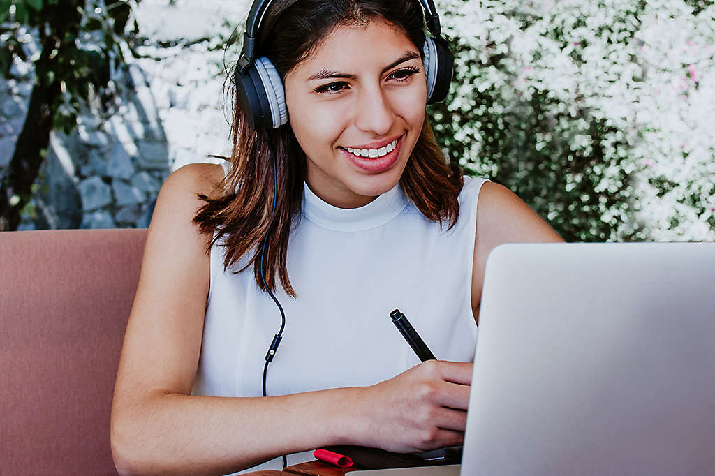 Woman smiling and taking notes while listening to a video on her laptop.