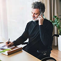 A man in a wheelchair sitting at a desk and talking on a cell phone. 