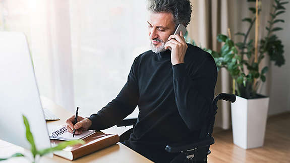 A man in a wheelchair sitting at a desk and talking on a cell phone. 