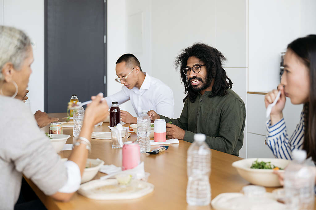 A group of employees sit in a breakroom eating packed lunches together.