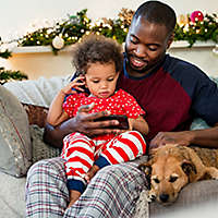 A father and daughter sitting on the couch with their dog, looking at his phone.