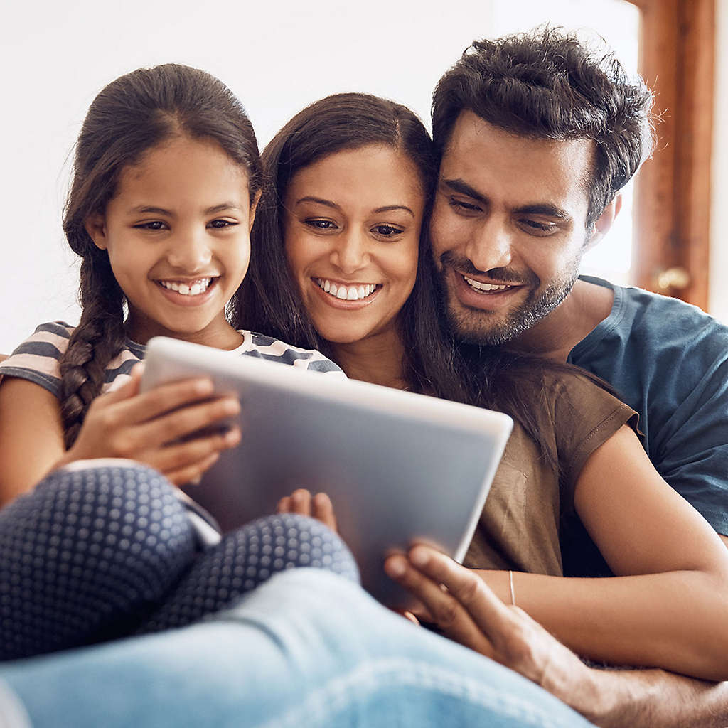A family sitting on the couch happily looking at something on a tablet together.