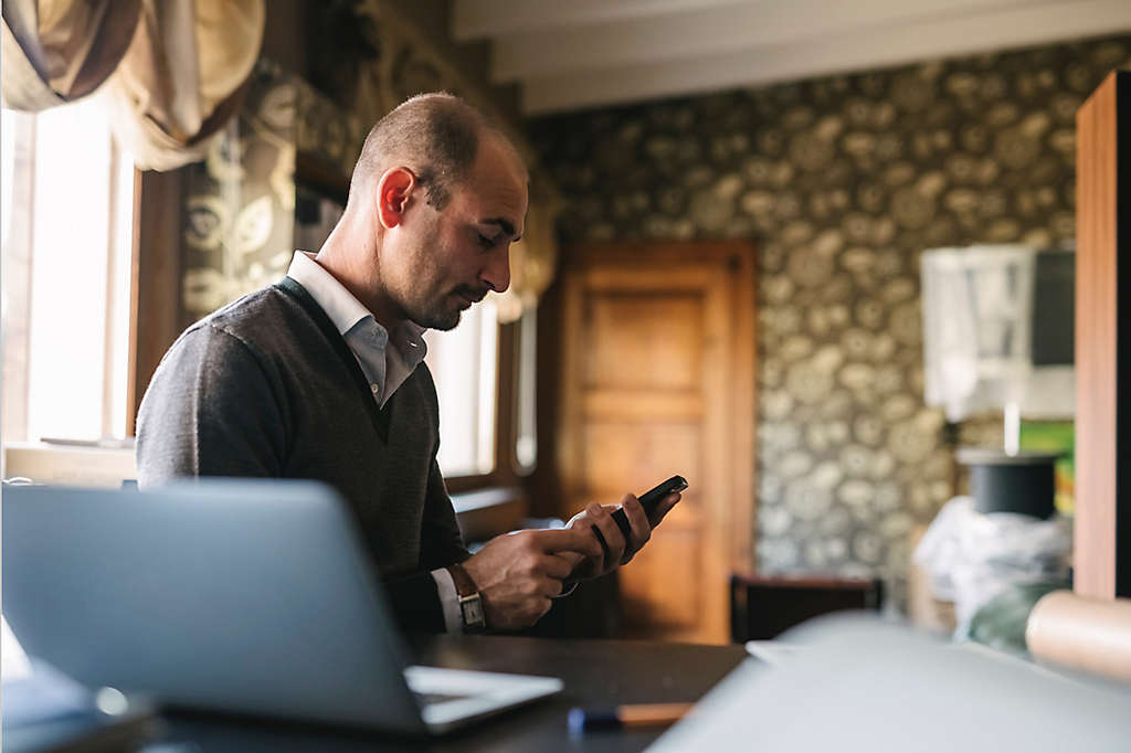 A man, sitting in his home office, checks the performance of investments on his phone.