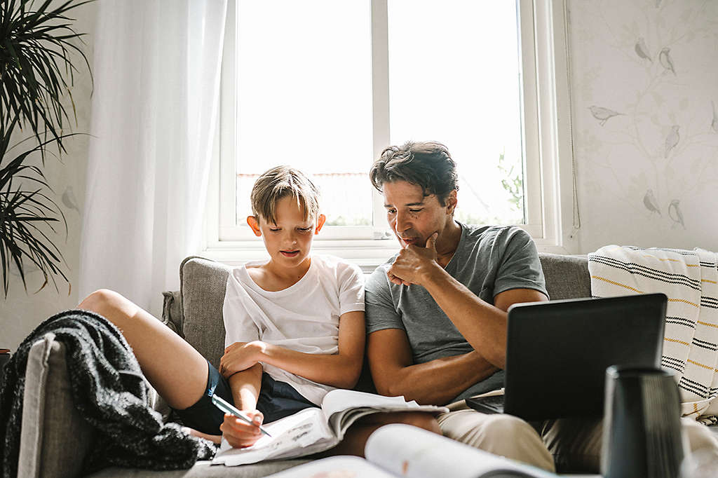 son looking in book while father sitting by with laptop on sofa at home