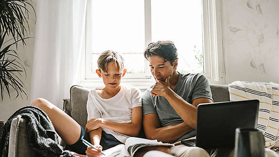 son looking in book while father sitting by with laptop on sofa at home