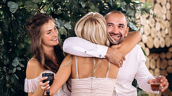Woman hugging couple while holding drinks and celebrating.