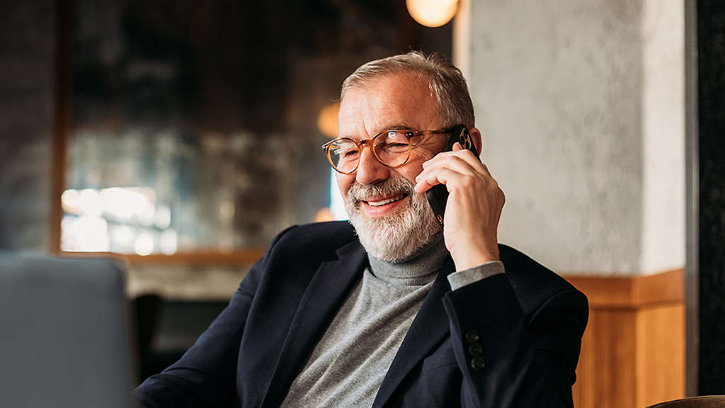 Older man with glasses smiling while talking on a smartphone in a cozy indoor setting.