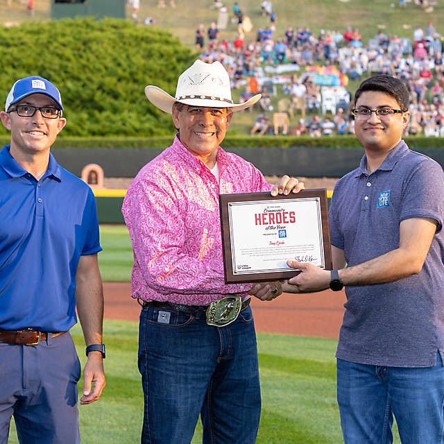Three men standing on a baseball field with a Community Heroes certificate