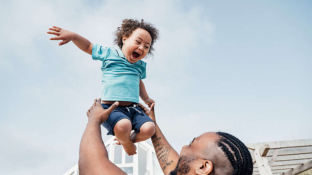 Over the shoulder view of 35 year old Black father lifting ecstatic 4 year old son high into the air as they enjoy outdoor playtime together.