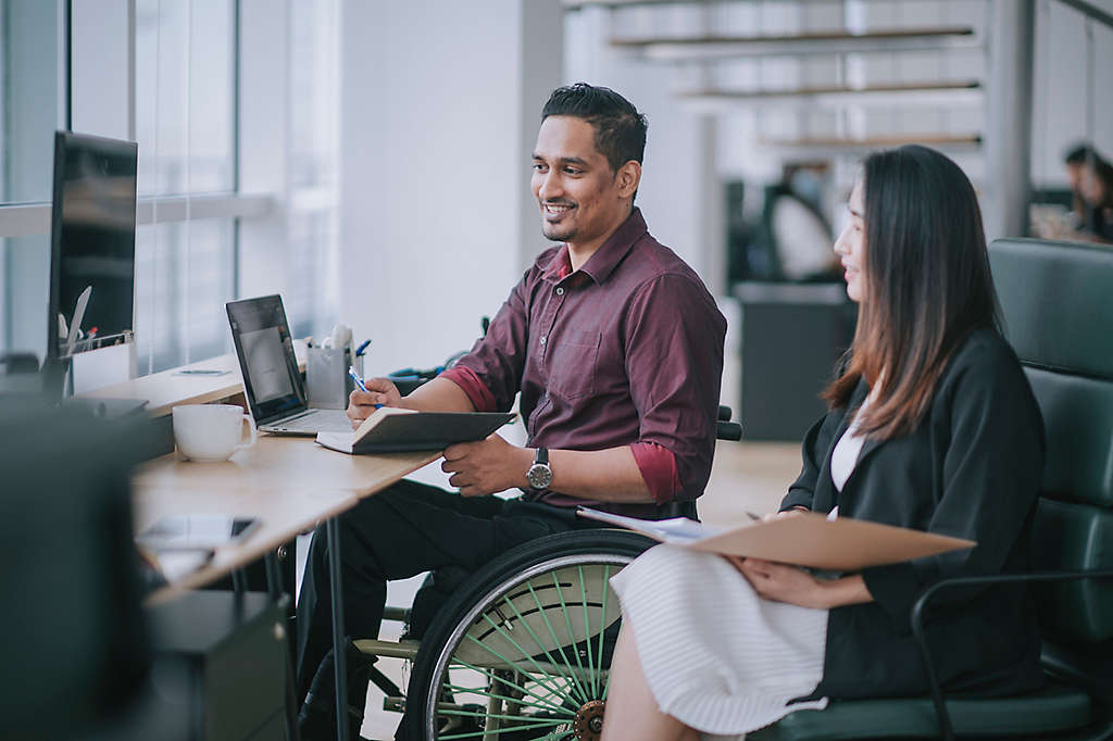 A white-collar male worker in a wheelchair has a conversation with his female colleague in the office 