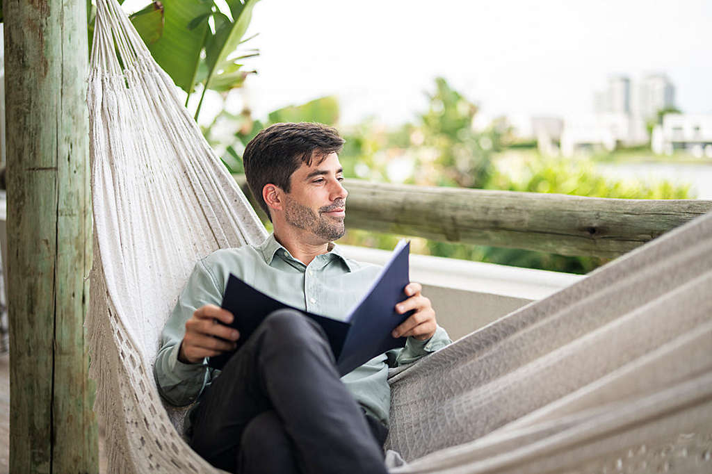Young man lying on a hammock while perusing work documents in his backyard at home