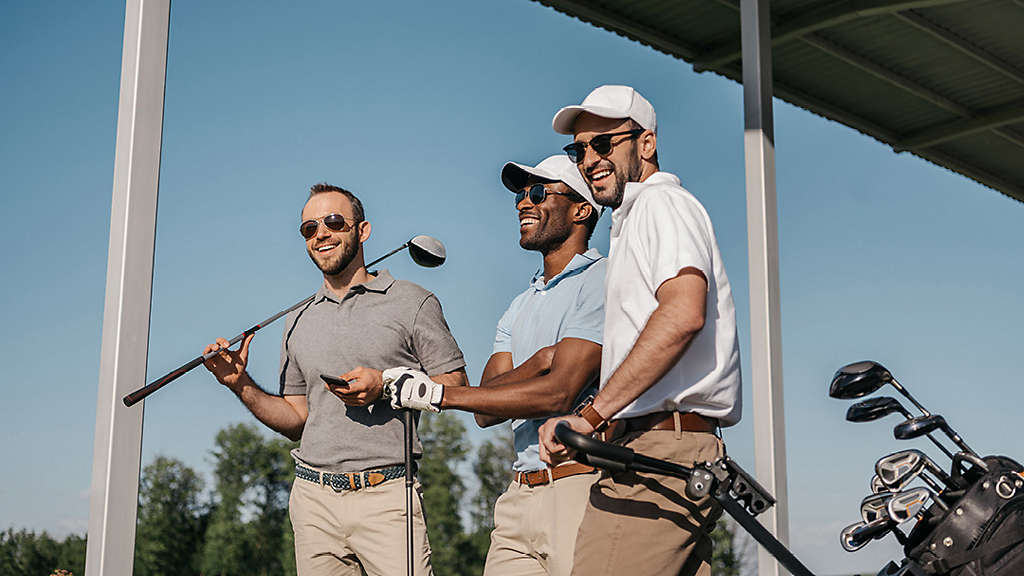 Three men smiling and standing together on a golf course, holding golf clubs and a golf bag nearby under a covered outdoor structure on a sunny day.