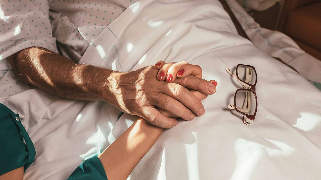 A daughter holds her father�s hand as he lies in a hospital bed.�