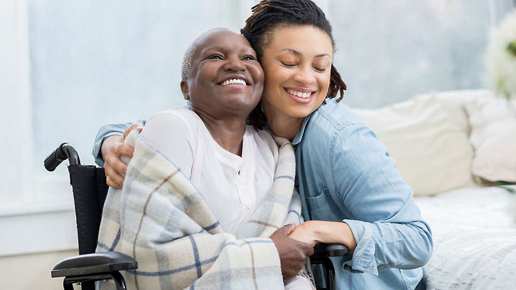 Granddaughter hugging grandmother in wheelchair and blanket in her home