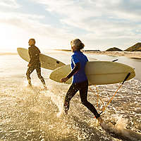 Two older people running into an ocean with surfboards